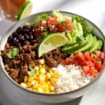 Ground beef burrito bowl with rice, black beans, corn, pico de gallo, lettuce, and sliced avocado, topped with a lime wedge.