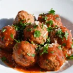Cooked ground beef meatballs coated in marinara sauce and topped with chopped parsley and grated Parmesan on a white plate.