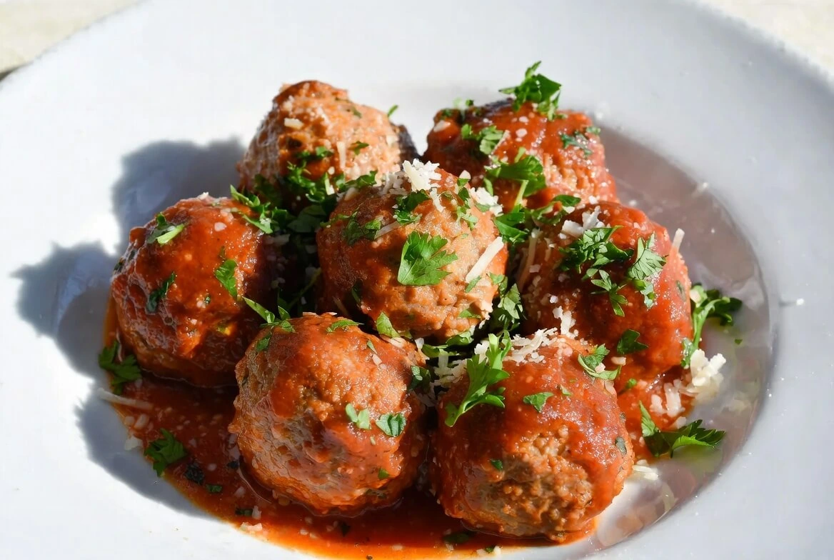 Cooked ground beef meatballs coated in marinara sauce and topped with chopped parsley and grated Parmesan on a white plate.