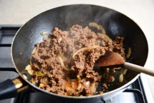 Ground beef cooking in a skillet with onions and spices, being stirred with a wooden spoon for burrito bowls.